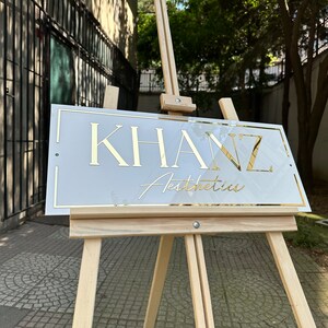 K&ouml;nnte beinhalten: Ein Schild f&uuml;r "Khanz Aesthetics" mit goldfarbenen Buchstaben und Zierleisten, das auf einer h&ouml;lzernen Staffelei ausgestellt ist. Das Schild hat einen wei&szlig;en Hintergrund und ist rechteckig. Die Staffelei ist aus hellem Holz gefertigt und befindet sich im Freien.