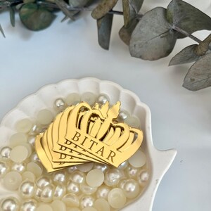 May include: Gold crown-shaped mirror acrylic ornaments with the name "BITAR" engraved on them. The ornaments are displayed in a white ceramic bowl filled with white pearl beads.
