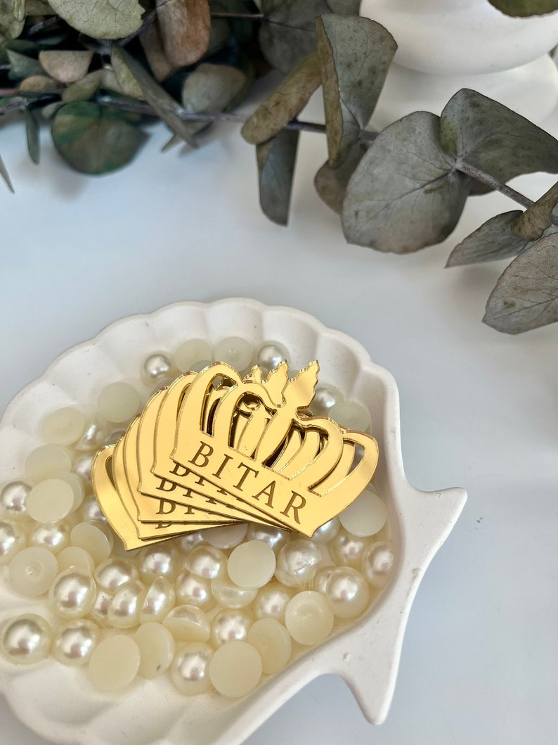 May include: Gold crown-shaped mirror acrylic ornaments with the name "BITAR" engraved on them. The ornaments are displayed in a white ceramic bowl filled with white pearl beads.