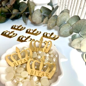 May include: Three gold crown-shaped decorations with the name "BITAR" inscribed on each. The crowns are sitting on a white seashell-shaped dish filled with white pearl-like beads.