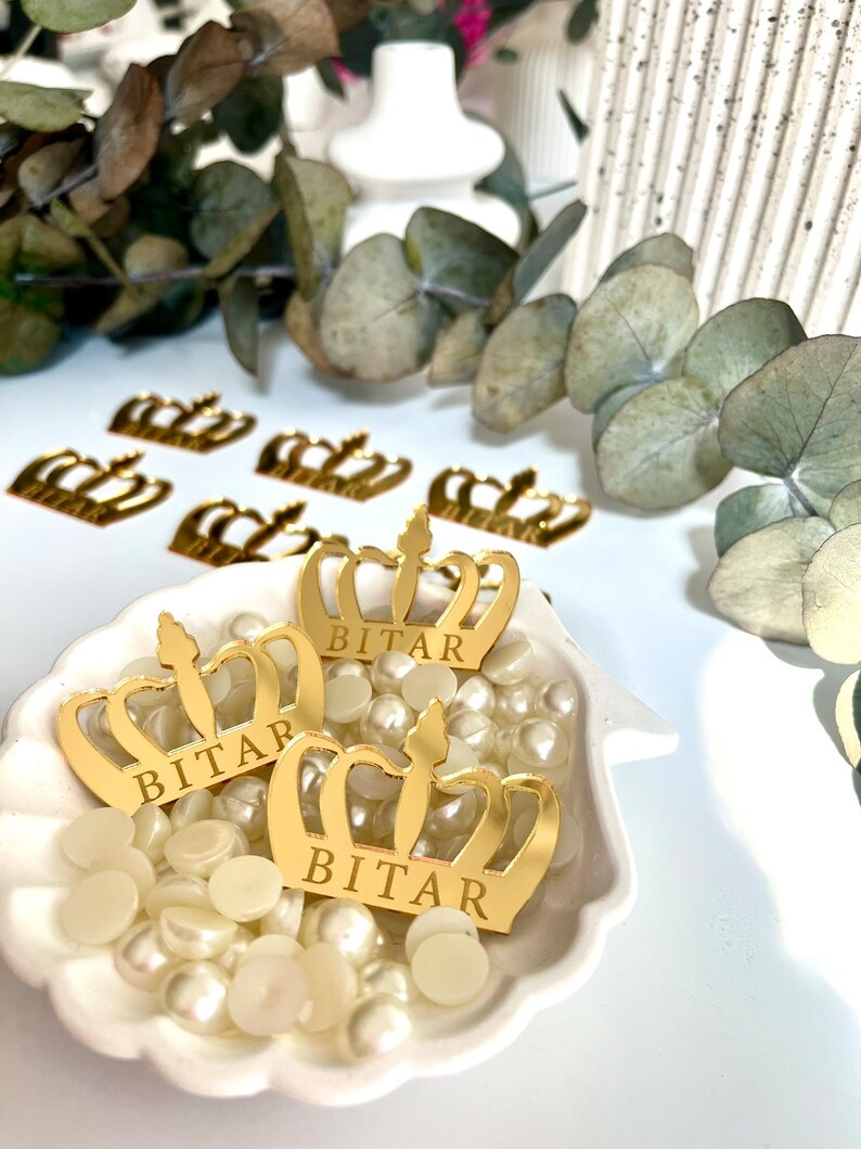 May include: Three gold crown-shaped decorations with the name "BITAR" inscribed on each. The crowns are sitting on a white seashell-shaped dish filled with white pearl-like beads.