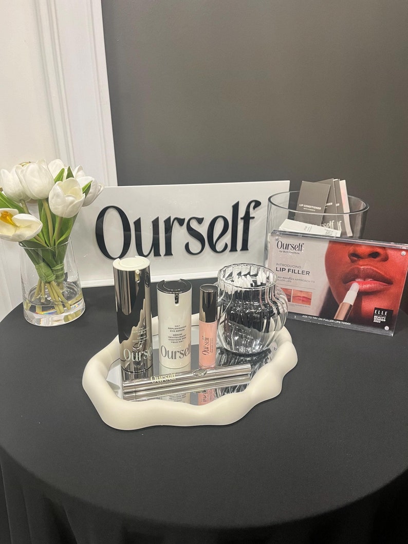 May include: A white tray with a silver mirrored surface displays a range of beauty products, including a lip filler, a serum, and a moisturiser. The products are all labelled "Ourself" and are presented on a black table with a white sign that reads "Ourself".