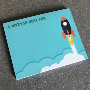 May include: A blue card with the text "A Mitzvah Note For" and a black and red rocket ship with a white flame trail.