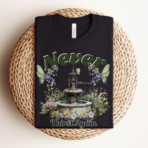 May include: Black t-shirt with a floral design featuring a fountain, butterflies, and the words "Never Thirst Again" and "John 4:14". The design includes green, white, and purple flowers.