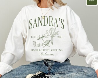 May include: White sweatshirt with a green graphic design that says "SANDRA'S EST. 2024 BACHELORETTE WEEKEND Bahamas". The graphic features a palm tree, beach chairs, and a cocktail.