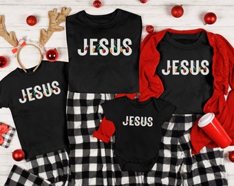 May include: A family matching set of black t-shirts with the word "JESUS" in white letters and Christmas lights. The shirts are worn by a family of four, including a baby, a child, a teenager, and an adult. The shirts are paired with black and white plaid pants.