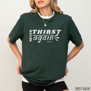 May include: Forest green t-shirt with the text "NEVER THIRST again" in white, with "John 4:14" below. The shirt has a white crew neck collar and is worn by a person.