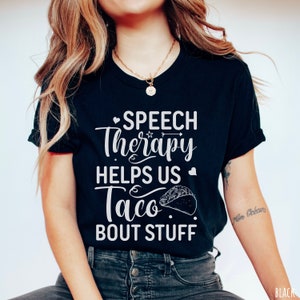 May include: Black t-shirt with white text that reads "Speech Therapy Helps Us Taco Bout Stuff". The text includes a heart and arrow graphic. A taco graphic is also included.
