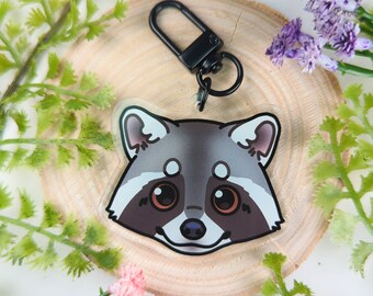 Puede incluir: Un llavero con un dije de cara de mapache de dibujos animados en blanco y negro con un anillo de llave de metal negro. El dije está hecho de acrílico transparente y tiene un acabado brillante.