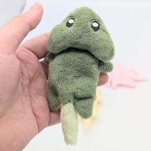 May include: A small, plush, olive-green creature toy with a soft, fuzzy texture. It features a wide, rounded head with two embroidered white eyes, short arms, and a lighter, pale green tail extending from the bottom of its rounded body.