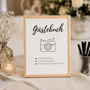 Könnte beinhalten: Ein rechteckiger Holzrahmen mit einem weißen Schild, das den schwarzen Schriftzug Gästebuch, ein Strichzeichnungs-Kamerasymbol mit Herzen sowie drei Punkte enthält: FOTO KNIPSEN, FOTO EINKLEBEN und NACHRICHT SCHREIBEN FÜR.