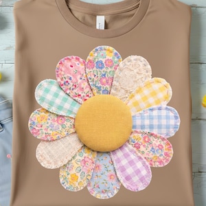 Könnte beinhalten: Ein hellbraunes T-Shirt mit einem großen aufgenähten Gänseblümchen-Design. Die Blütenblätter bestehen aus verschiedenen gemusterten Stoffen in Pastellfarben mit einem gelben Zentrum. Hellblaue Jeans mit floraler Stickerei sind ebenfalls zu sehen.