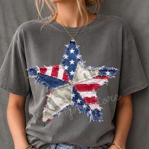 May include: A gray t-shirt featuring a large star design made from patches of the American flag and camouflage fabric. The star has a distressed, frayed edge. The shirt is a relaxed fit and has short sleeves.