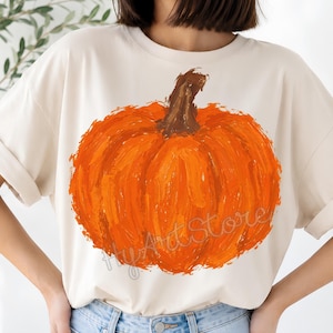 Peut inclure: Un t-shirt à manches courtes de couleur crème arborant un grand motif de citrouille orange peint avec une tige brune. Le design présente des textures de coups de pinceau visibles et le texte HyArtStore est imprimé en écriture grise sur la citrouille.