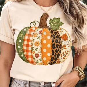 Op de afbeelding: Een crèmekleurig T-shirt met korte mouwen en een grote, centrale afbeelding van een pompoen. De pompoen is opgebouwd uit verticale segmenten met diverse patronen, waaronder oranje ruitjes, bloemenprints, witte stippen en luipaardprint.