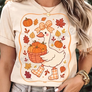 May include: Cream-coloured t-shirt with a cartoon goose carrying a basket of pumpkins. The goose wears orange and white plaid boots and a bow. The design includes orange pumpkins, autumn leaves, and a decorative border.