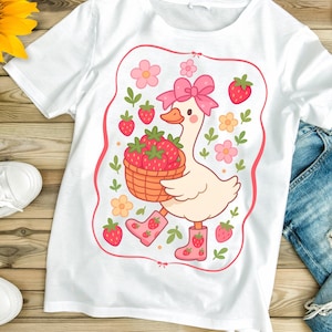K&ouml;nnte beinhalten: Wei&szlig;es T-Shirt mit einer Cartoon-Illustration eines Entchens, das eine rosa Schleife tr&auml;gt und einen Korb mit Erdbeeren tr&auml;gt. Das Entchen ist von rosa Blumen und gr&uuml;nen Bl&auml;ttern umgeben.