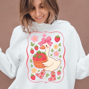 K&ouml;nnte beinhalten: Wei&szlig;er Pullover mit einem rosa und wei&szlig;en Cartoon-Entchen, das einen Korb mit Erdbeeren tr&auml;gt. Das Entchen tr&auml;gt rosa Stiefel und eine rosa Schleife. Das Design ist von einem rosa Rahmen mit wei&szlig;en Blumen und Erdbeeren umgeben.