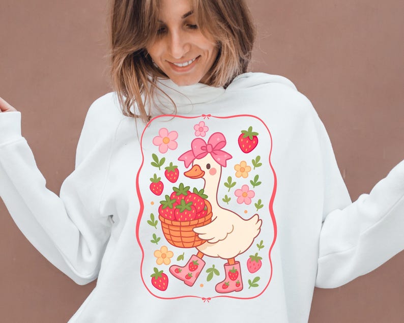 K&ouml;nnte beinhalten: Wei&szlig;er Pullover mit einem rosa und wei&szlig;en Cartoon-Entchen, das einen Korb mit Erdbeeren tr&auml;gt. Das Entchen tr&auml;gt rosa Stiefel und eine rosa Schleife. Das Design ist von einem rosa Rahmen mit wei&szlig;en Blumen und Erdbeeren umgeben.