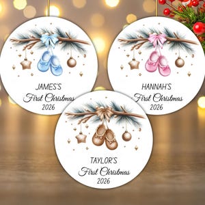 Pode incluir: Três ornamentos redondos brancos com ilustrações de ramos de pinheiro, laços e sapatos de bebé em azul, rosa e castanho. Cada ornamento apresenta o texto First Christmas 2026 com os nomes James's, Hannah's ou Taylor's impressos acima.