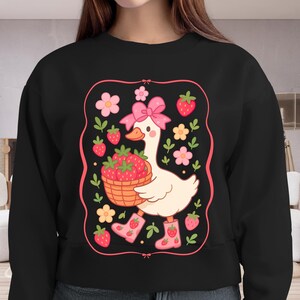 K&ouml;nnte beinhalten: Schwarzer Pullover mit einer wei&szlig;en Gans, die einen Korb mit Erdbeeren h&auml;lt. Die Gans tr&auml;gt eine rosa Schleife und rosa Stiefel. Das Design ist von rosa Blumen und Erdbeeren umgeben.