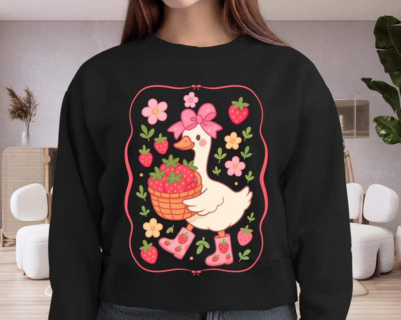 K&ouml;nnte beinhalten: Schwarzer Pullover mit einer wei&szlig;en Gans, die einen Korb mit Erdbeeren h&auml;lt. Die Gans tr&auml;gt eine rosa Schleife und rosa Stiefel. Das Design ist von rosa Blumen und Erdbeeren umgeben.