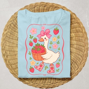 K&ouml;nnte beinhalten: Ein hellblaues T-Shirt mit einer Cartoon-Illustration einer Gans, die eine rosa Schleife tr&auml;gt und einen Korb mit Erdbeeren tr&auml;gt. Die Gans ist von Blumen und Erdbeeren umgeben.