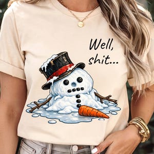 May include: A cream-colored t-shirt featuring a graphic of a melting snowman wearing a black top hat with a red band. The snowman has stick arms, a carrot nose, and coal-like buttons. The text "Well, shit..." is printed in black above the snowman.