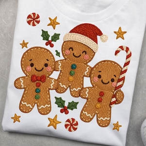 May include: White t-shirt featuring an embroidered design of three gingerbread men. One wears a Santa hat and another holds a candy cane. The design includes stars, holly, and candy accents, creating a festive holiday theme.