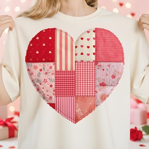 Puede incluir: Camiseta color crema con un gran diseño de corazón de patchwork. El corazón está compuesto por varias telas rojas y rosas con patrones como lunares, rayas y estampados florales. Una prenda casual para el día a día.