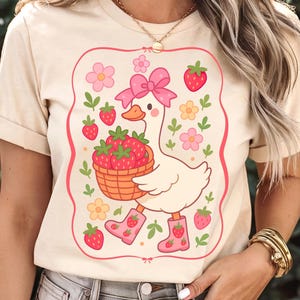 K&ouml;nnte beinhalten: Ein wei&szlig;es T-Shirt mit einer Cartoon-Illustration einer Gans, die rosa Stiefel tr&auml;gt und einen Korb mit Erdbeeren tr&auml;gt. Die Gans ist von rosa Blumen und gr&uuml;nen Bl&auml;ttern umgeben. Der Text "Strawberry Goose" ist auf das Shirt gedruckt.