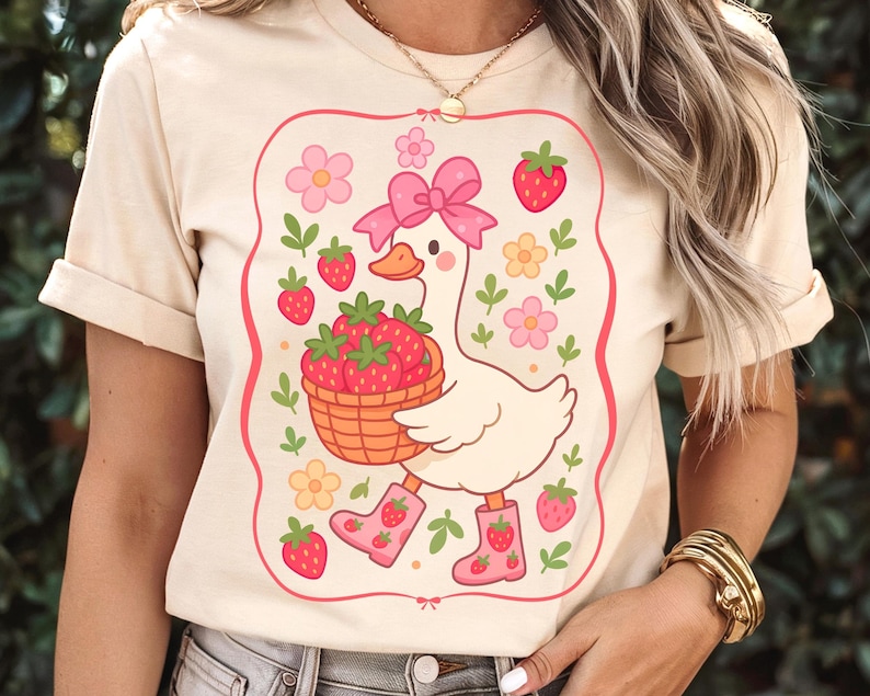 K&ouml;nnte beinhalten: Ein wei&szlig;es T-Shirt mit einer Cartoon-Illustration einer Gans, die rosa Stiefel tr&auml;gt und einen Korb mit Erdbeeren tr&auml;gt. Die Gans ist von rosa Blumen und gr&uuml;nen Bl&auml;ttern umgeben. Der Text "Strawberry Goose" ist auf das Shirt gedruckt.