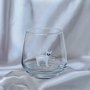May include: A clear glass tumbler with a white glass cat figurine inside. The cat has blue eyes and a red nose.