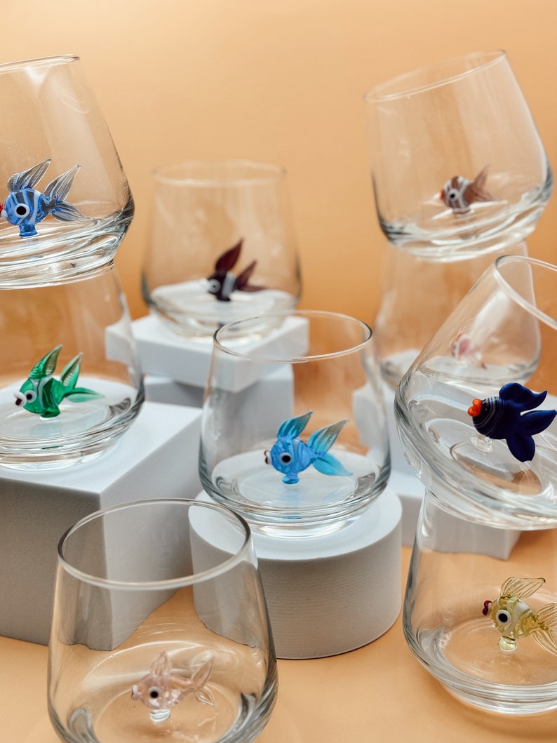 May include: A collection of clear glass tumblers with colorful glass fish figurines inside. The tumblers are arranged on white square pedestals and a light brown surface.