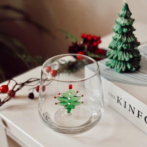 Puede incluir: Un vaso transparente con un dise&ntilde;o de &aacute;rbol de Navidad verde y rojo en su interior. El vaso est&aacute; sobre una superficie blanca con un libro titulado "The Kinfolk" en el fondo.