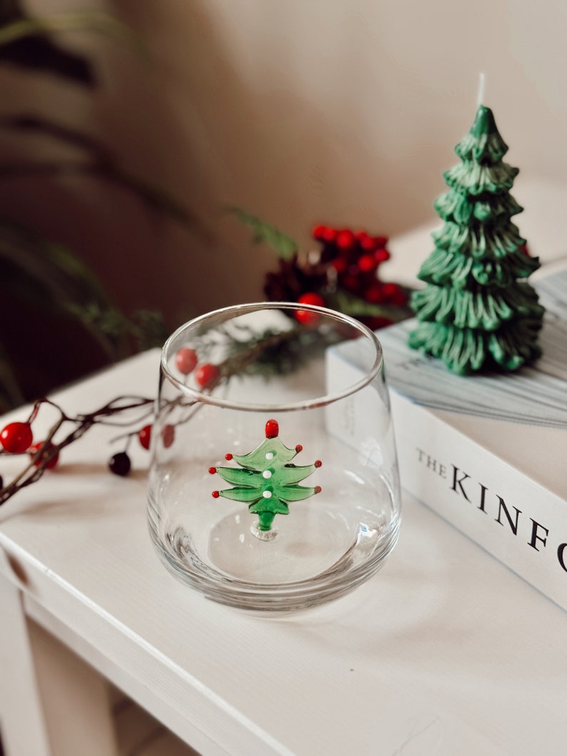 Puede incluir: Un vaso transparente con un dise&ntilde;o de &aacute;rbol de Navidad verde y rojo en su interior. El vaso est&aacute; sobre una superficie blanca con un libro titulado "The Kinfolk" en el fondo.