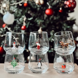 Puede incluir: Tres vasos de vidrio transparente con un dise&ntilde;o de doble capa. Cada vaso presenta una figura diferente con tem&aacute;tica navide&ntilde;a en su interior: un mu&ntilde;eco de nieve, un ping&uuml;ino y un reno. Los vasos est&aacute;n dispuestos sobre una superficie blanca frente a un &aacute;rbol de Navidad desenfocado.
