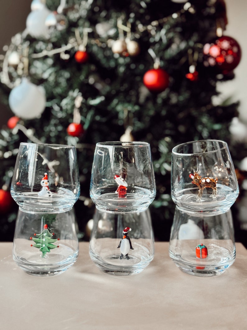 Puede incluir: Tres vasos de vidrio transparente con un dise&ntilde;o de doble capa. Cada vaso presenta una figura diferente con tem&aacute;tica navide&ntilde;a en su interior: un mu&ntilde;eco de nieve, un ping&uuml;ino y un reno. Los vasos est&aacute;n dispuestos sobre una superficie blanca frente a un &aacute;rbol de Navidad desenfocado.
