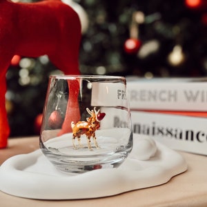 Puede incluir: Un vaso transparente con una peque&ntilde;a figura de reno dorada en su interior. El vaso est&aacute; sobre un posavasos blanco en forma de nube. El vaso est&aacute; delante de una pila de libros con el texto "French Window" y "Renaissance" visible.