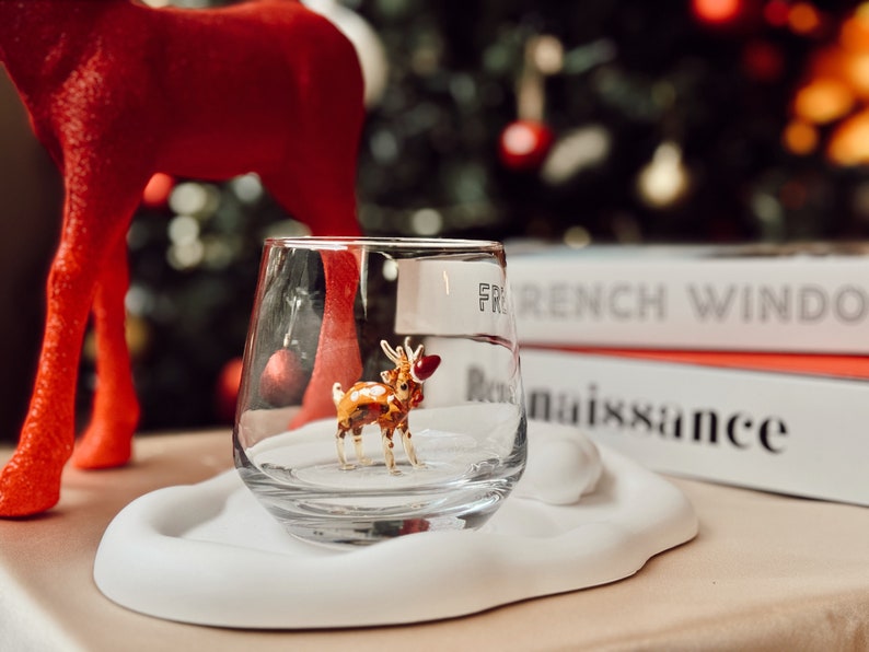 Puede incluir: Un vaso transparente con una peque&ntilde;a figura de reno dorada en su interior. El vaso est&aacute; sobre un posavasos blanco en forma de nube. El vaso est&aacute; delante de una pila de libros con el texto "French Window" y "Renaissance" visible.