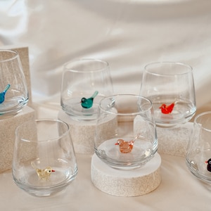 May include: Six clear glass tumblers with different colored glass birds inside. The glasses are sitting on white pedestals.