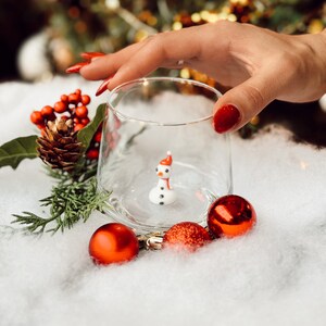 Puede incluir: Una peque&ntilde;a figura de mu&ntilde;eco de nieve blanco con un sombrero rojo dentro de un vaso transparente. El vaso est&aacute; sobre una superficie blanca y esponjosa con adornos navide&ntilde;os rojos.