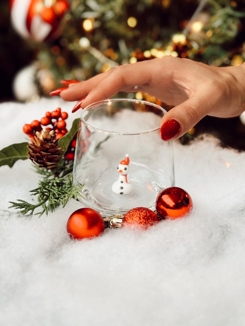 Puede incluir: Una peque&ntilde;a figura de mu&ntilde;eco de nieve blanco con un sombrero rojo dentro de un vaso transparente. El vaso est&aacute; sobre una superficie blanca y esponjosa con adornos navide&ntilde;os rojos.