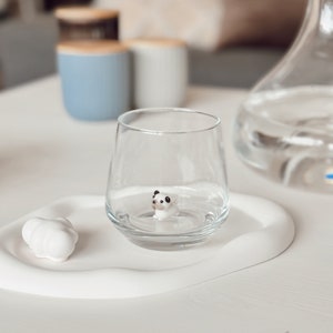 May include: A white ceramic coaster with a small white figurine of a panda bear inside a clear glass. The coaster is on a white table.