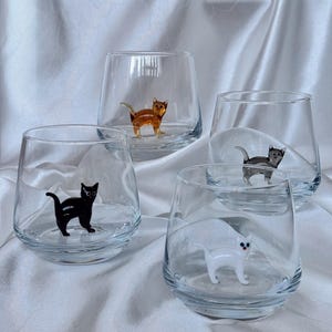 May include: Four clear glass tumblers each featuring a small, colorful cat figurine inside. The cats are in various colors: orange, black, gray, and white. The glasses are set against a white, textured background.