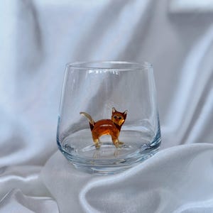 May include: A clear glass with a brown glass cat figurine inside. The glass is sitting on a white fabric surface.