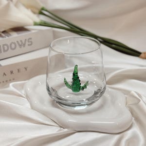 May include: A clear glass with a green alligator figurine inside, resting on a white, organically shaped ceramic plate. The glass and plate sit on a white satin surface. Decorative white flowers and a book are in the background.