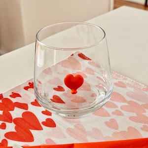 May include: A clear, stemless glass with a red heart-shaped accent. The glass sits on a heart-patterned napkin. The napkin has a white background with red hearts. The glass is empty.