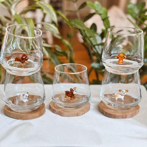 May include: Clear stemless glasses with small, colorful dog figurines inside, resting on wooden coasters. The dogs are various breeds and colors. The glasses are arranged on a white surface, with greenery in the background.
