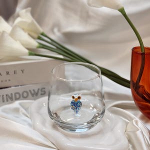 May include: A clear glass tumbler with a small, colourful owl figurine inside, resting on a white, organically shaped ceramic plate. The background includes white calla lilies, a red vase, and a book.